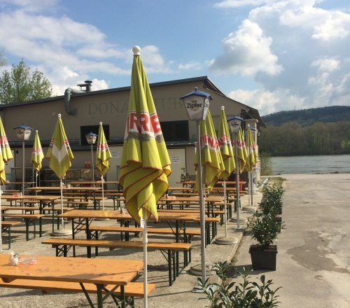 Biergarten mit gelben Sonnenschirmen am Flussufer, bewölkter Himmel., © donaustubn